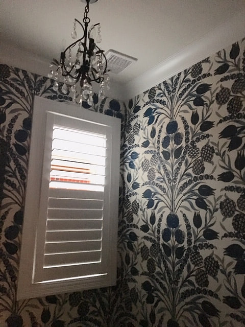 A bathroom papered in a navy and cream botanical print, with a shuttered window and a chandelier